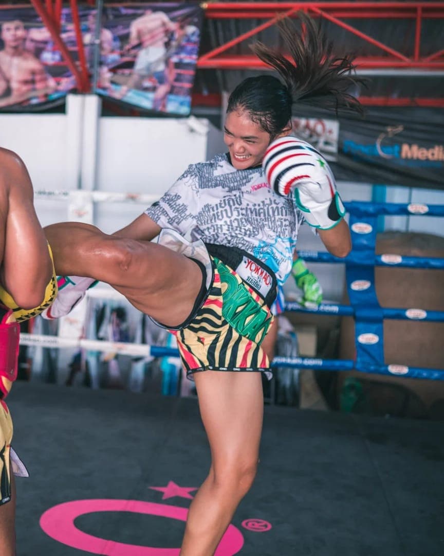 Ashley S. smiling as she lands a kick on pads in a Muay Thai ring