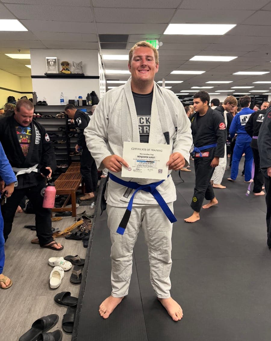 Ben H. in a white gi and blue belt, holding his Brazilian Jiu-Jitsu belt certificate